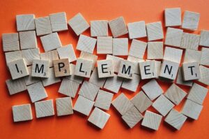 Wooden letter tiles spelling "IMPLEMENT" scattered among blank tiles on an orange background. 「IMPLEMENT」と綴られた木製の文字タイルが、無地のタイルとともにオレンジ色の背景の上に散らばっている。