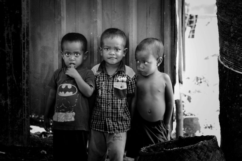 Three young children standing side by side in a modest environment, their serious expressions capturing attention. 質素な環境の中で並んで立つ3人の幼い子どもたち。真剣な表情が印象的。
